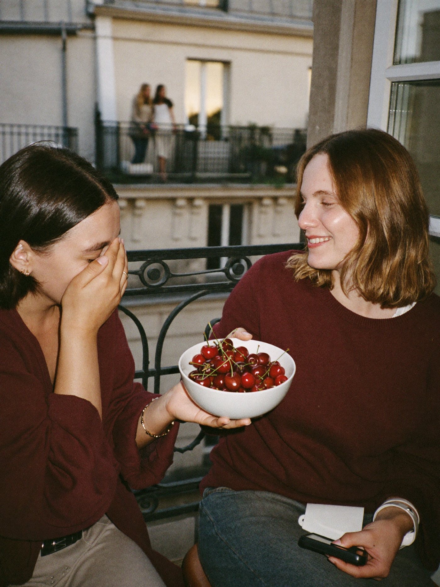 Deux amies rient sur un balcon parisien en partageant des cerises