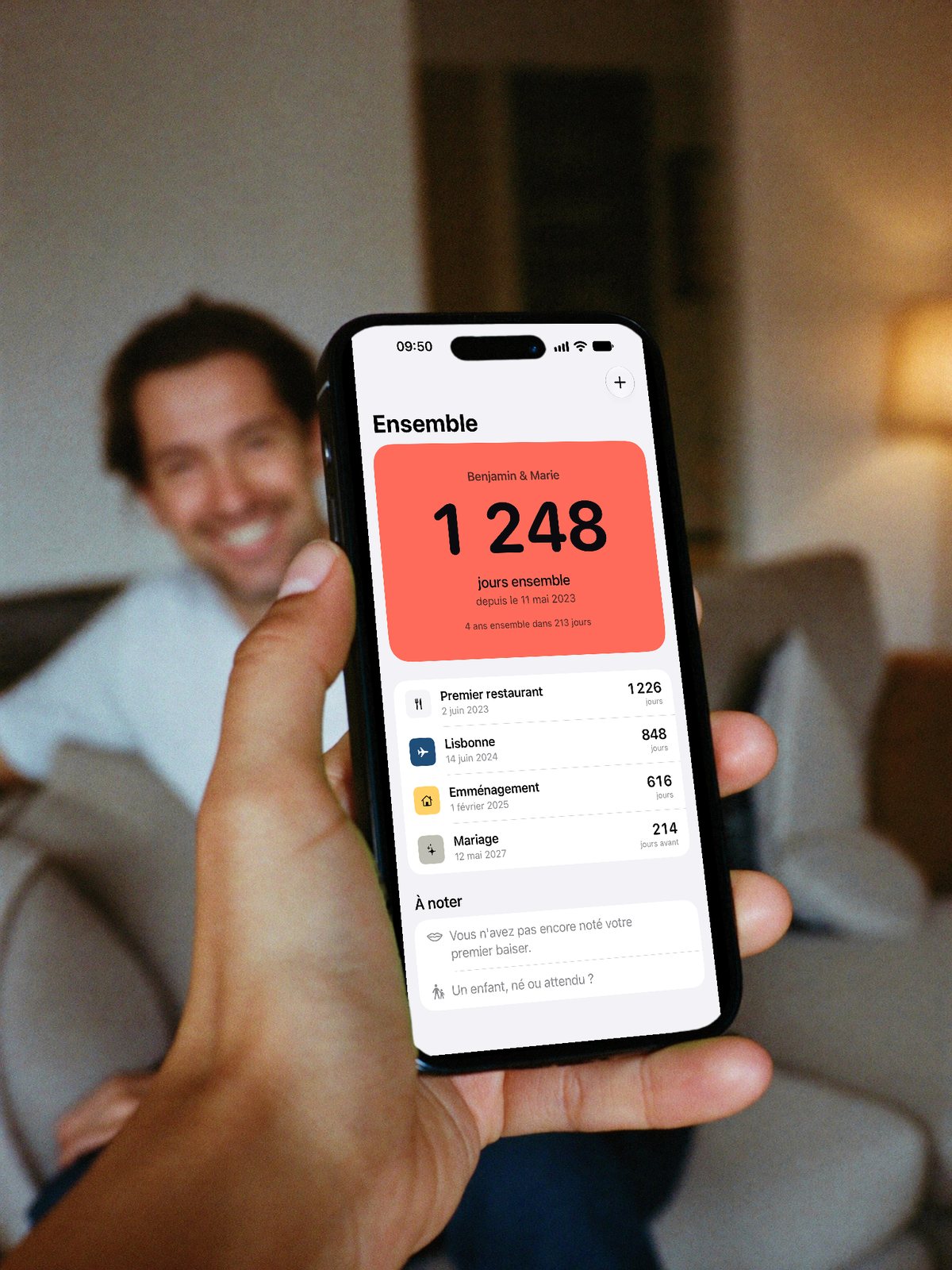 Une main tient un iPhone qui affiche Ensemble ; au fond, un homme sourit sur le canapé
